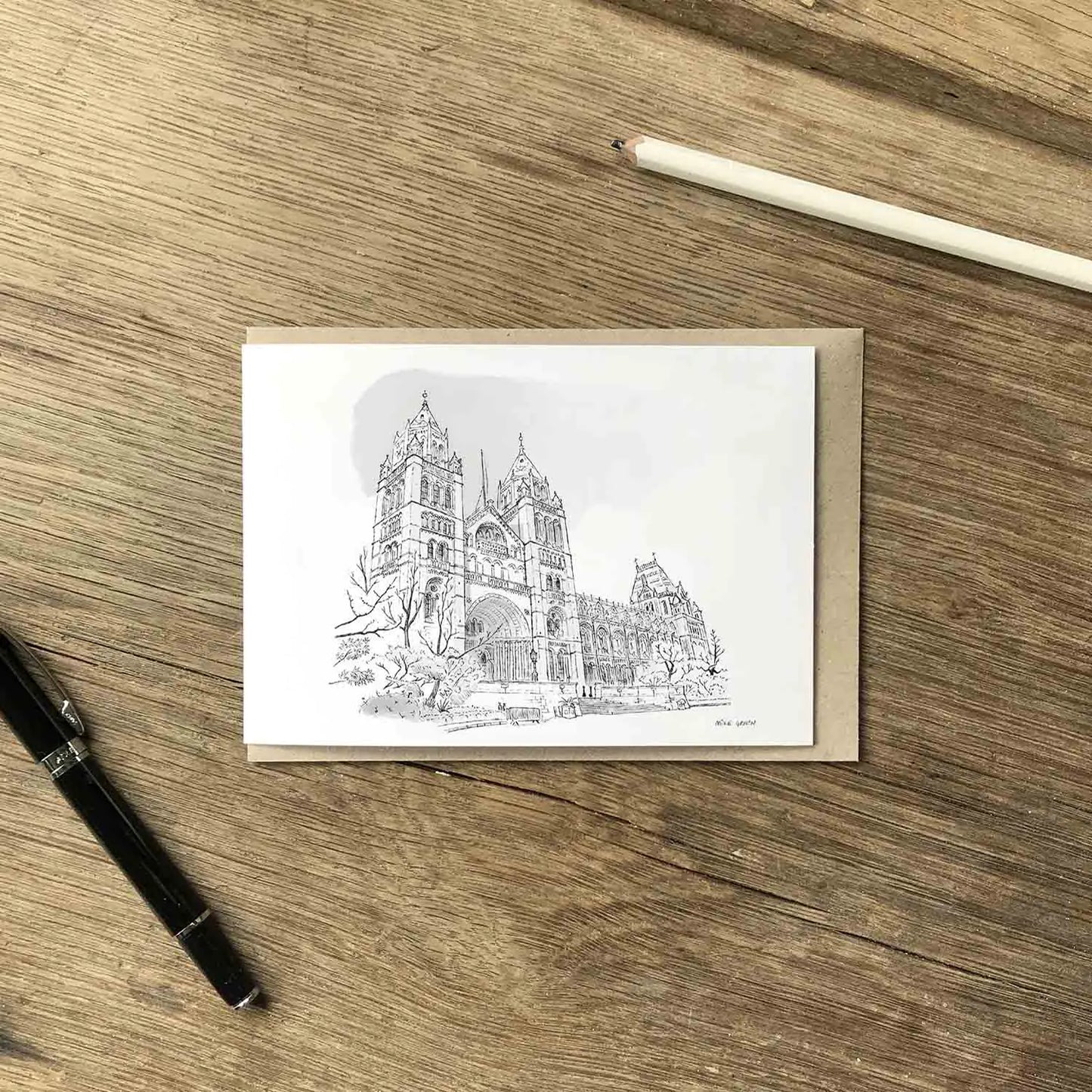 A white greeting card featuring a sketch of London's Natural History Museum in Kensington, displayed on a brown kraft envelope over a wooden desk with a white pencil, and a black pen.