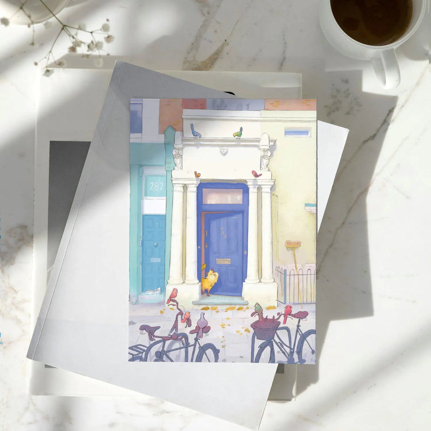 A greetings card with Notting Hill's Blue door from the film with a cat and birds in London next to a cup of coffee
