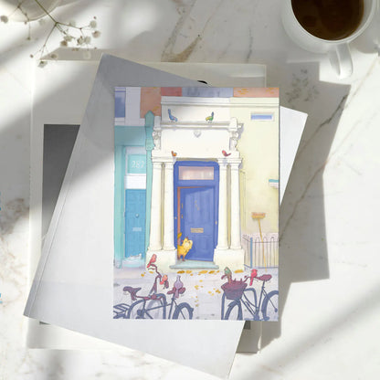 A greetings card with Notting Hill's Blue door from the film with a cat and birds in London next to a cup of coffee