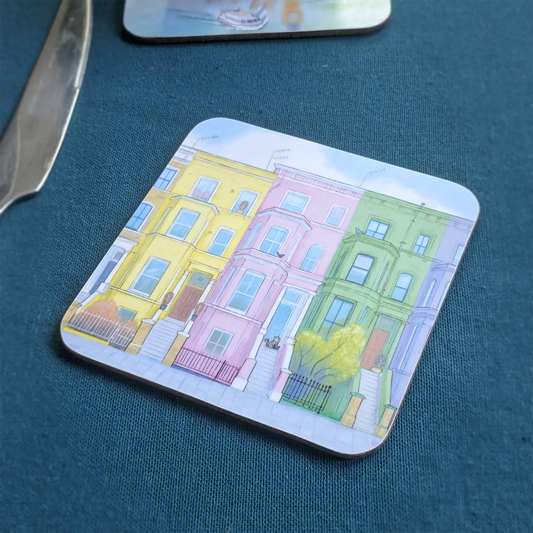 A square coaster featuring an illustration of colorful houses in London's Notting Hill with a cat, placed on a textured dark teal surface next to a knife.