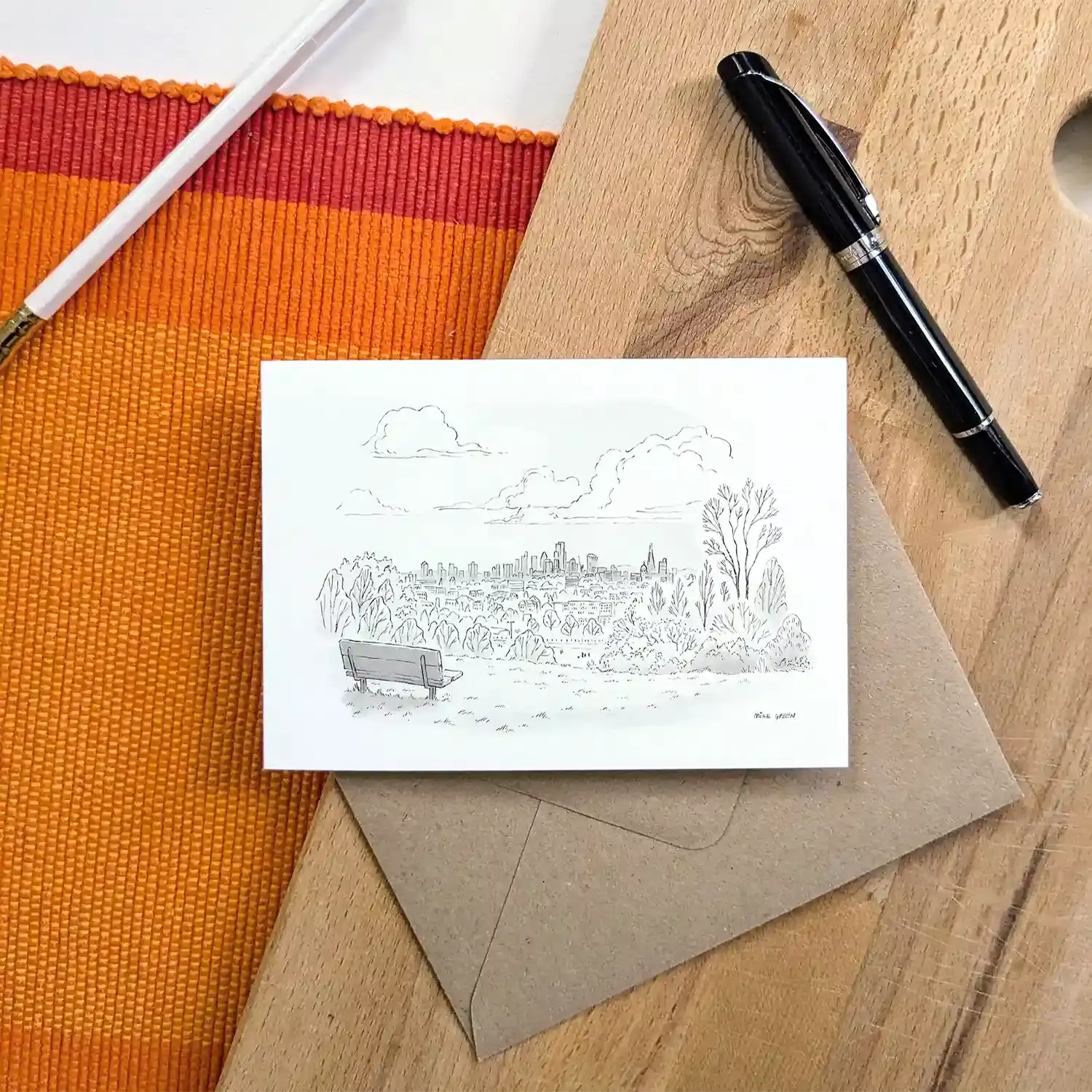 A greeting card featuring a black ink sketch of Hampstead Heath in north London, placed on top of a brown kraft envelope on a wooden surface with an orange striped mat, a white pencil, and a black pen as props.