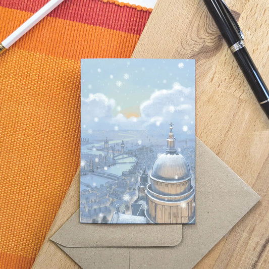 Christmas greetings card with London's St Pauls Cathedral in the snow at nighttime shown on a wooden surface with a pencil and pen