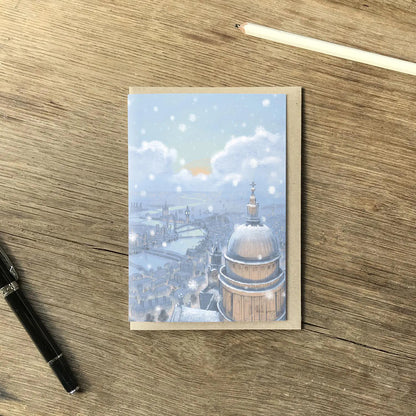 Christmas greetings card with London's St Pauls Cathedral in the snow at nighttime shown on a wooden surface with a pencil and pen