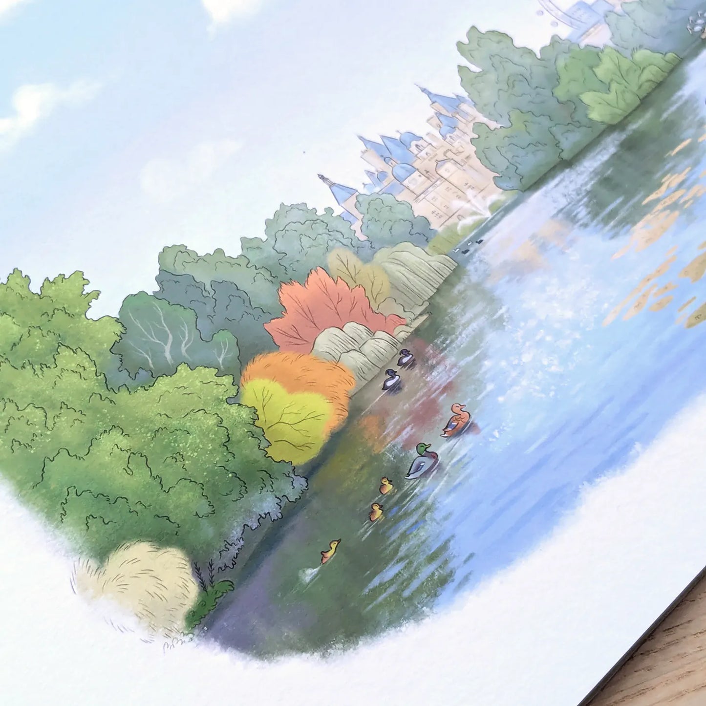 A watercolor-style illustration of St James Park lake in London with ducks and pelicans. The wall art print is shown at an angle on a white paper background with a small portion of a wooden surface visible at the bottom right.
