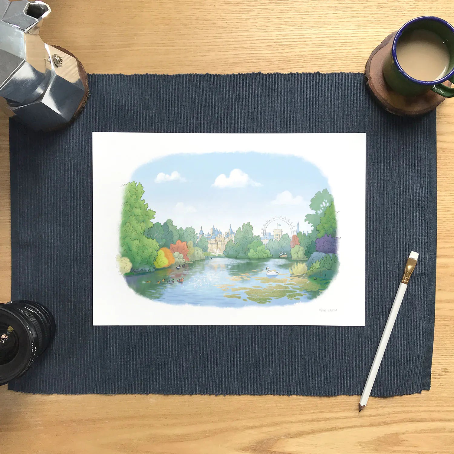 A watercolor landscape wall art print of St-James's Park in London showing the lake, wildlife and the London eye in the background. The print is displayed on a dark blue textured placemat on a wooden desk with coffee and drawing accessories.