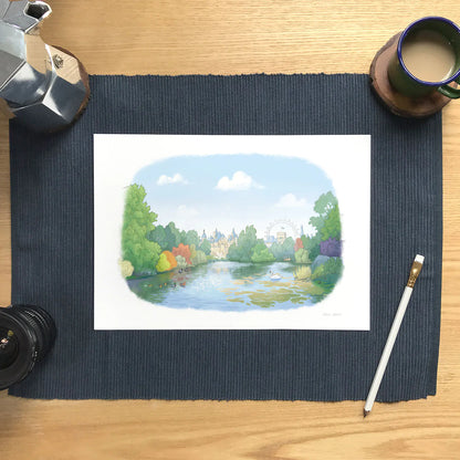 A watercolor landscape wall art print of St-James's Park in London showing the lake, wildlife and the London eye in the background. The print is displayed on a dark blue textured placemat on a wooden desk with coffee and drawing accessories.
