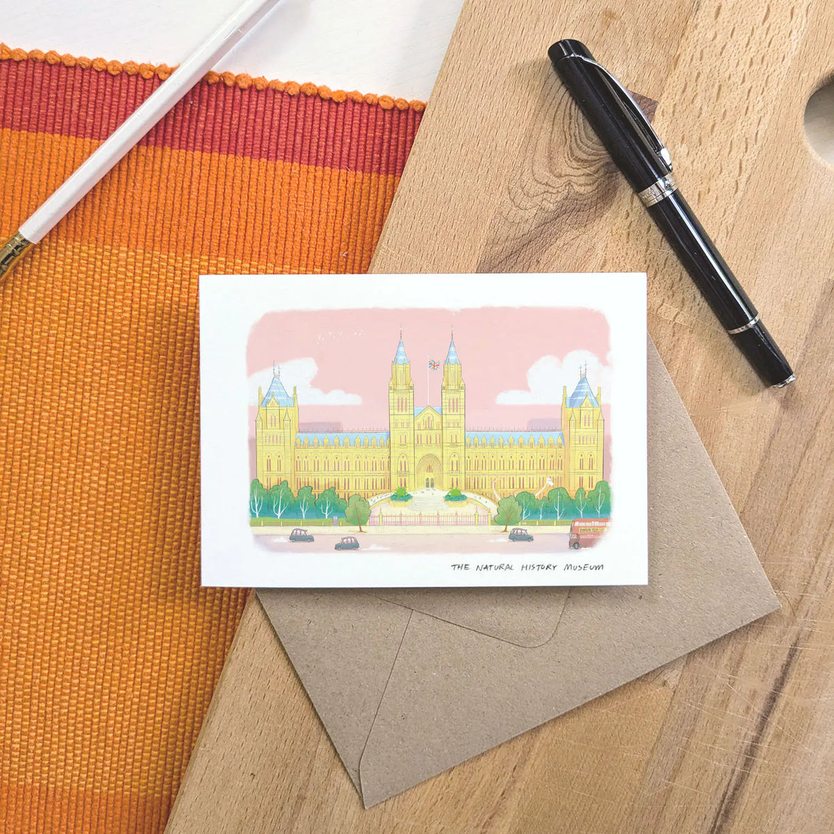 A greetings card featuring a pastel colour illustration of The Natural History Museum in London, layered over a brown kraft envelope on a wooden surface with a pen and pencil