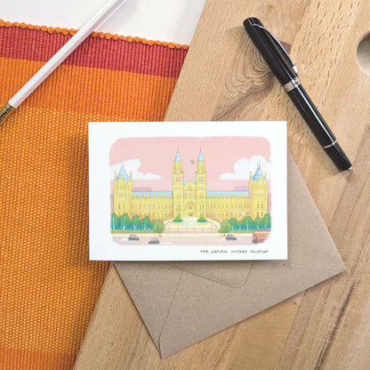 A greetings card featuring a pastel colour illustration of The Natural History Museum in London, layered over a brown kraft envelope on a wooden surface with a pen and pencil