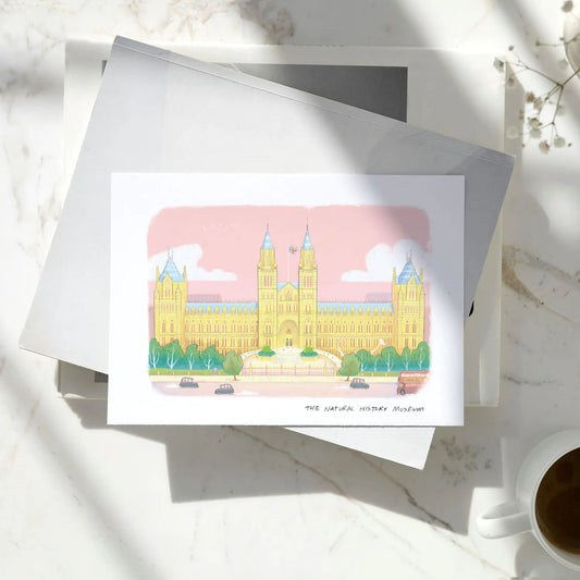 A greetings card featuring a pastel colour illustration of The Natural History Museum in London, layered over a brown kraft envelope on a pile of books