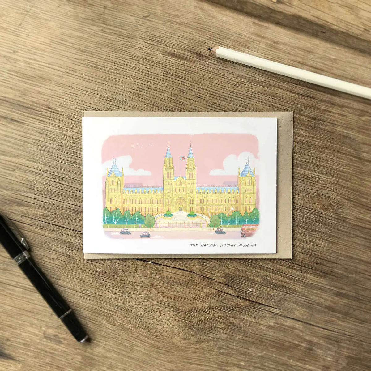 A greetings card featuring a pastel colour illustration of The Natural History Museum in London, layered over a brown kraft envelope on a wooden surface with a pen and pencil