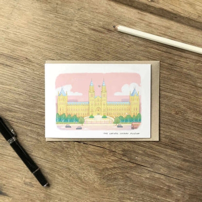 A greetings card featuring a pastel colour illustration of The Natural History Museum in London, layered over a brown kraft envelope on a wooden surface with a pen and pencil