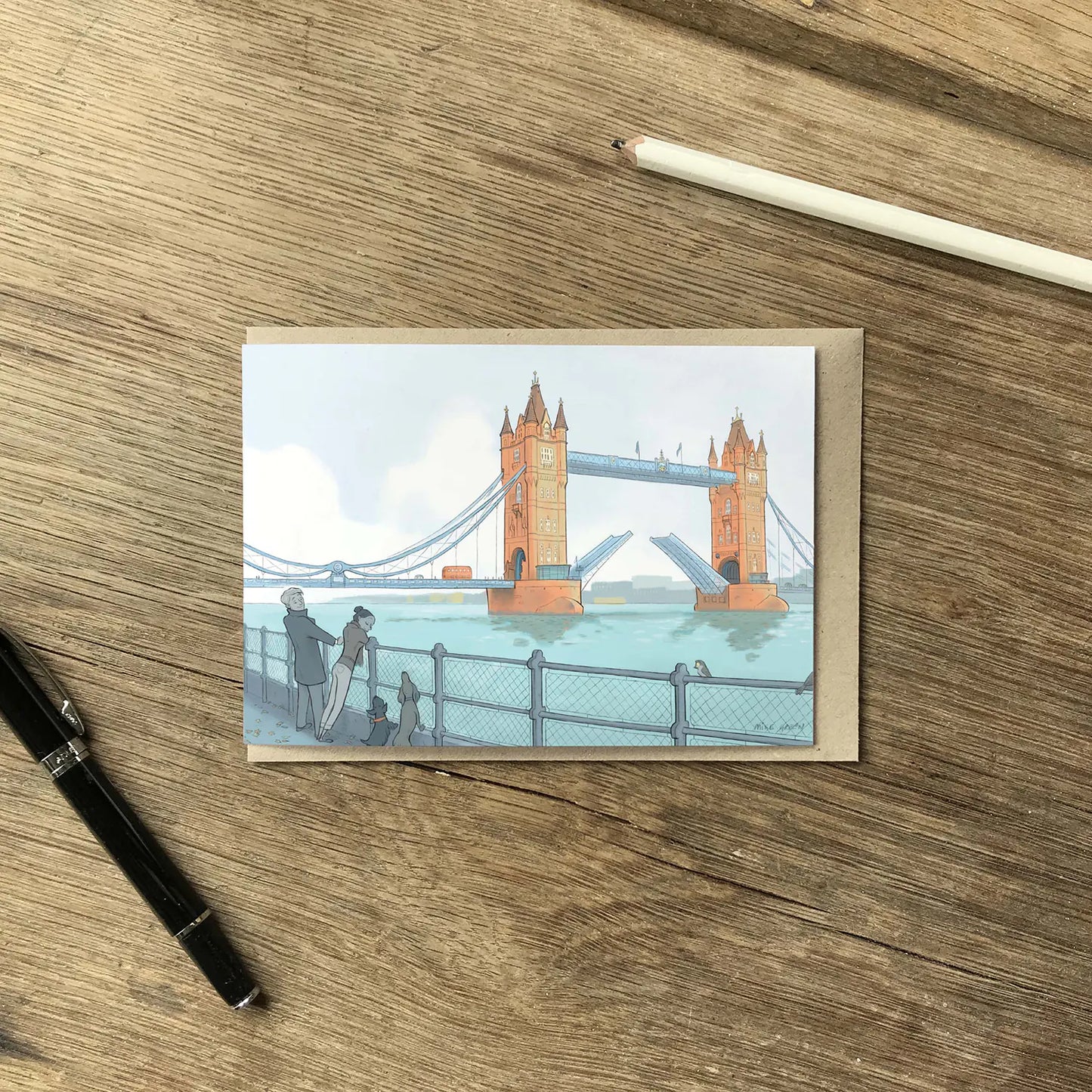 London's Tower Bridge with dog walkers illustrated on a greeting card with a pencil and pen on a wooden surface.