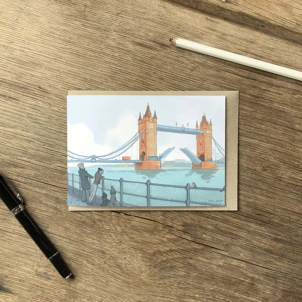 London's Tower Bridge with dog walkers illustrated on a greeting card with a pencil and pen on a wooden surface.