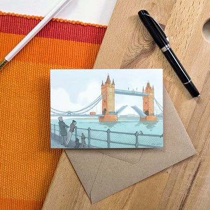 London's Tower Bridge with dog walkers illustrated on a greeting card with a pencil and pen on a wooden surface.