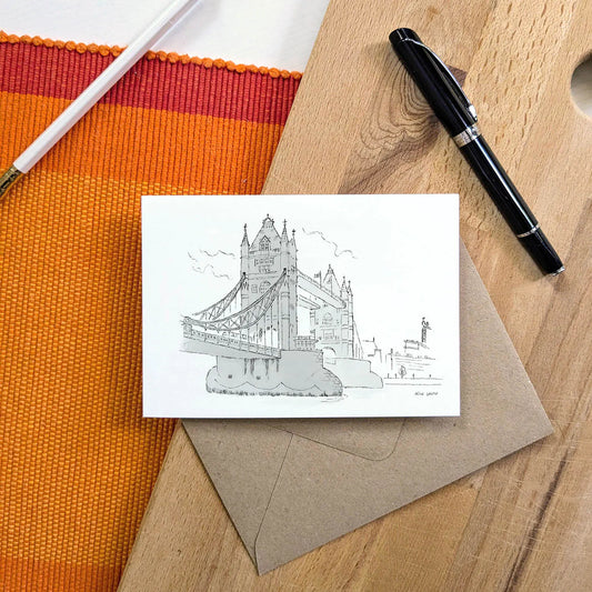 A greeting card featuring a black line drawing of Tower Bridge, on a wooden tabletop with a brown envelope, orange woven mat, white pencil, and black pen.
