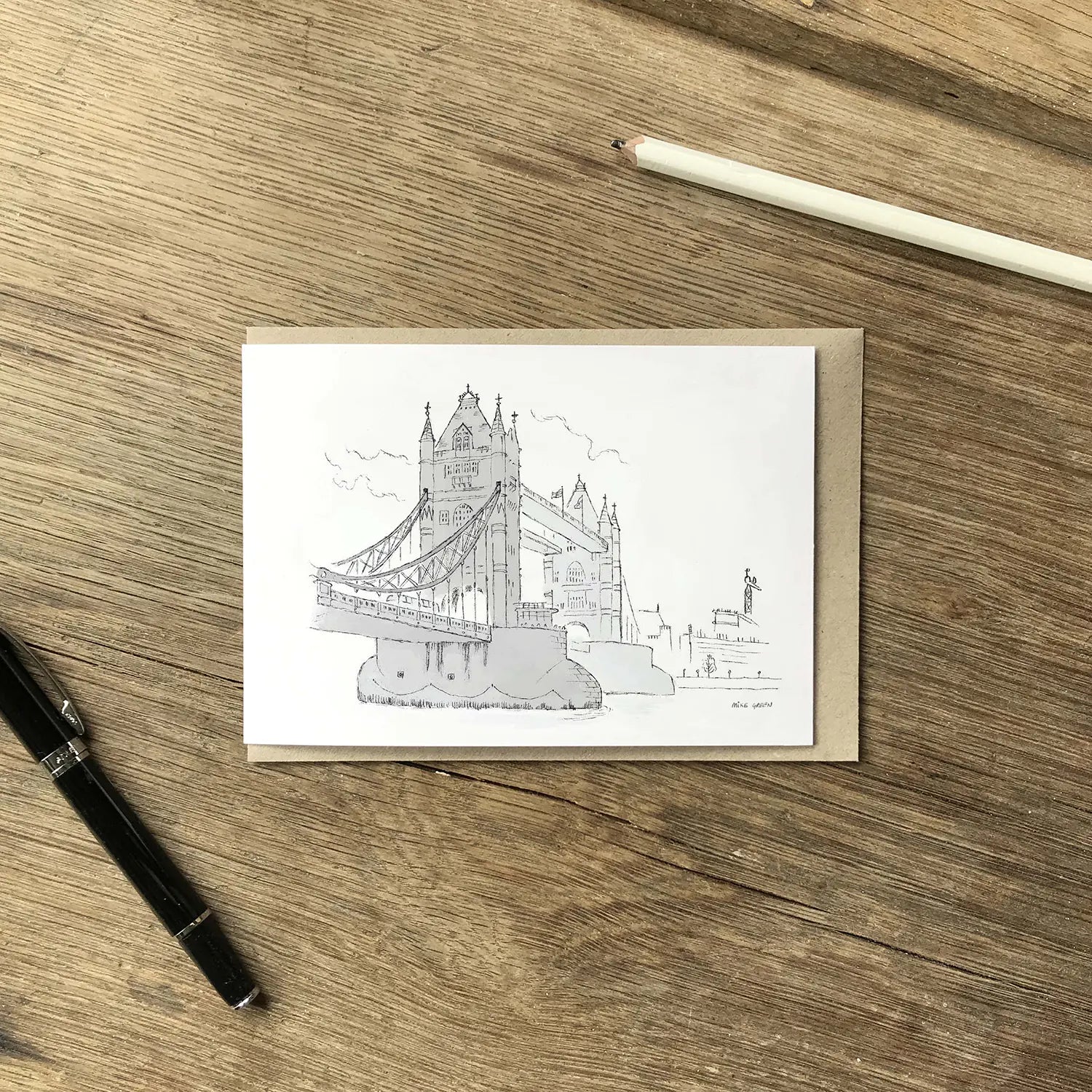 A greeting card featuring a black line drawing of Tower Bridge, on a wooden tabletop with a brown envelope, white pencil, and black pen.