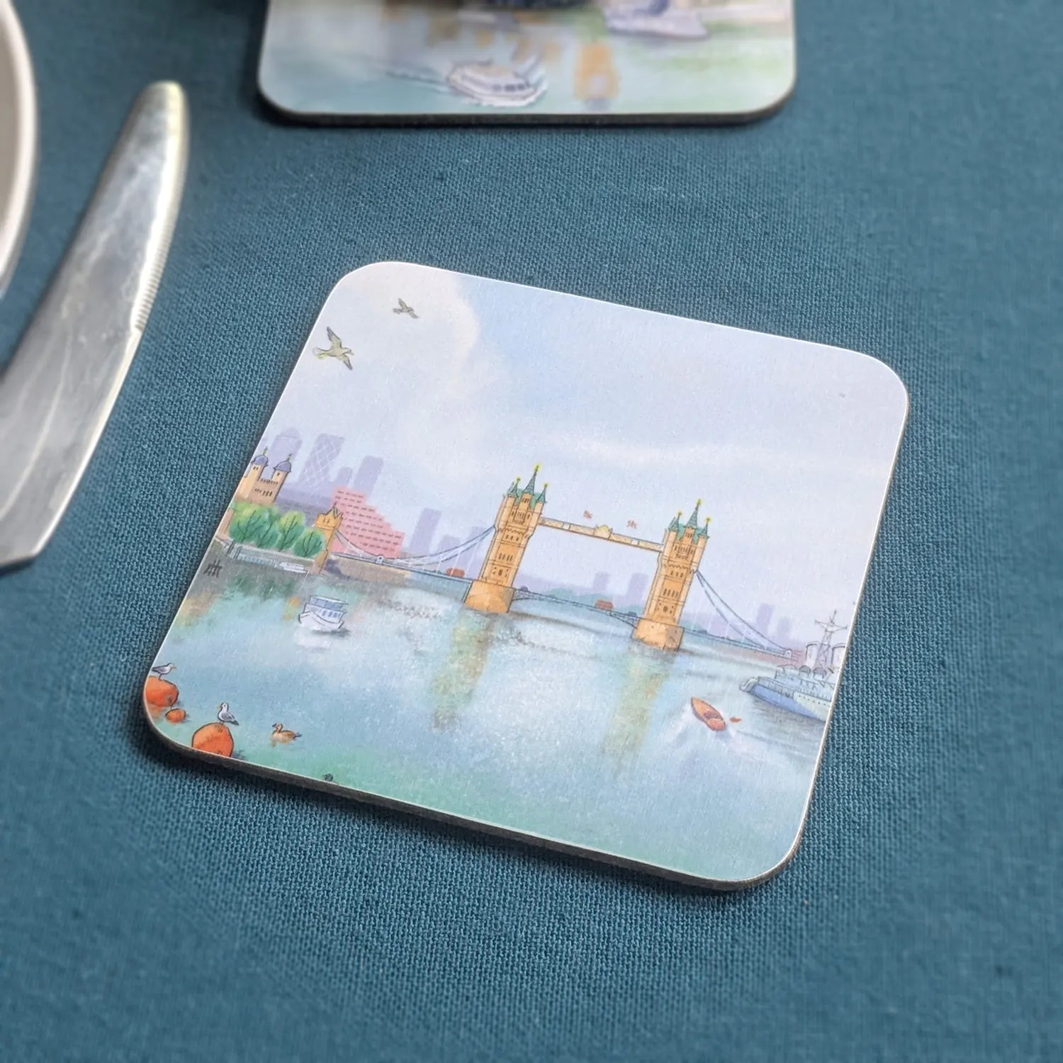 A square coasters with rounded corners featuring a watercolor illustration of London's Tower Bridge, placed on a textured dark teal table next to a silver knife.