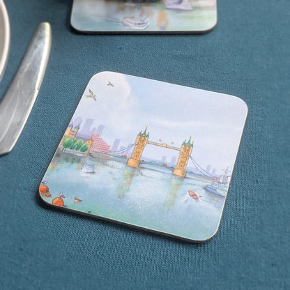 A square coasters with rounded corners featuring a watercolor illustration of London's Tower Bridge, placed on a textured dark teal table next to a silver knife.
