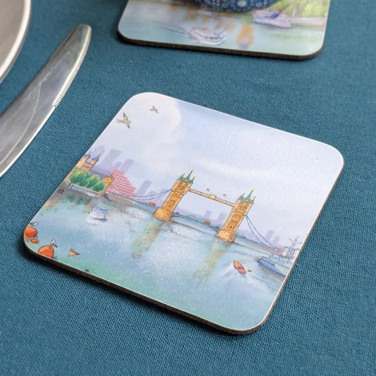 A square coaster with rounded corners featuring an Illustration of Tower Bridge in London, placed on a textured teal-blue table next to a knife. Another coaster is partially visible at the top.
