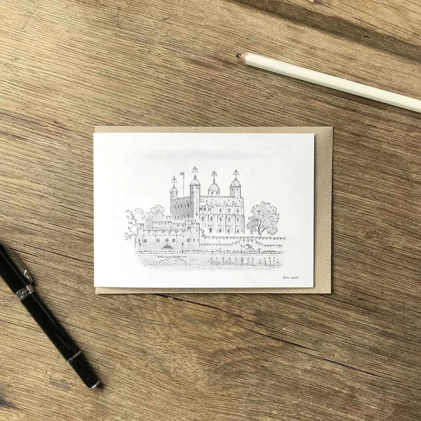 The Tower of London illustrated on a white greeting card with a brown kraft envelope, black pen and white pencil on a wooden surface
