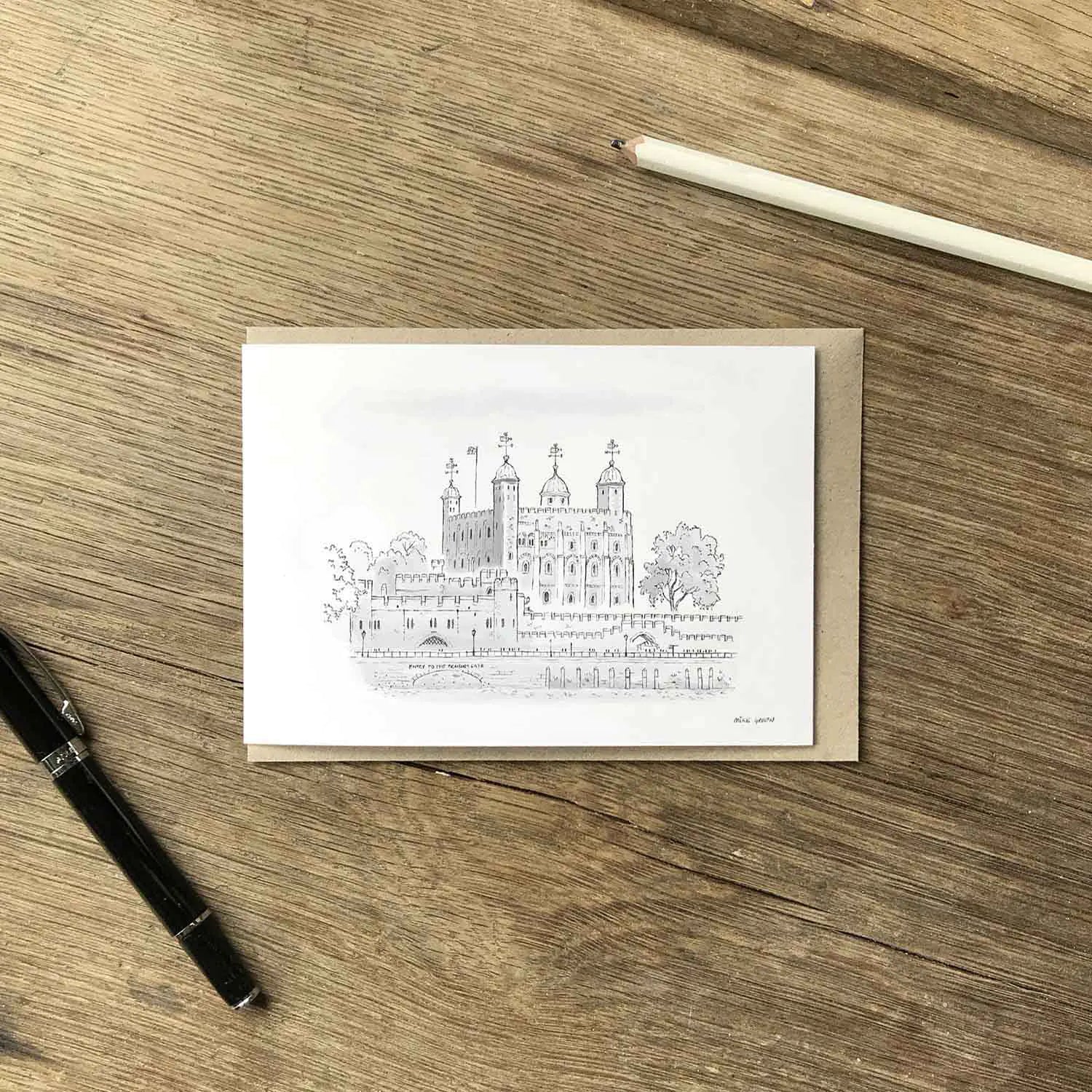 The Tower of London illustrated on a white greeting card with a brown kraft envelope, black pen and white pencil on a wooden surface