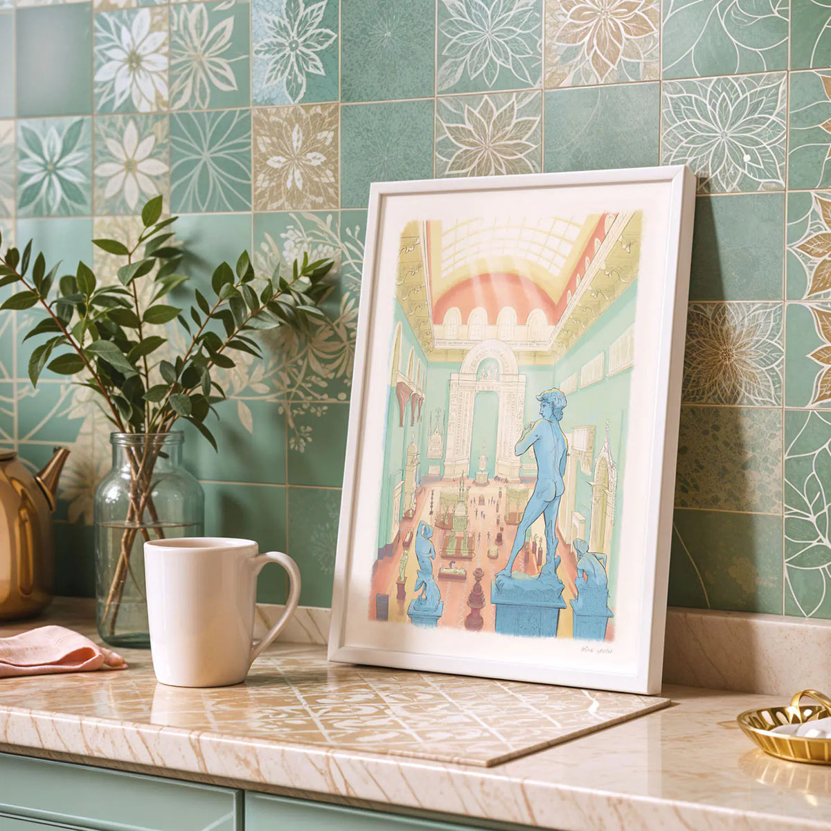 A framed art print of the Victoria and Albert Museum's Weston Cast Court in London's South Kensington leaning against a green patterned tile wall on a marble countertop, styled with a white mug, a glass vase with greenery, and a gold dish.