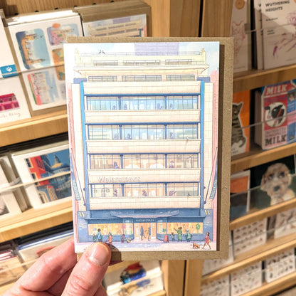 A hand holds a greetings card with featuring a painting of Waterstones Piccadilly bookshop in the shop, set against a blurred background of a book or card shop.