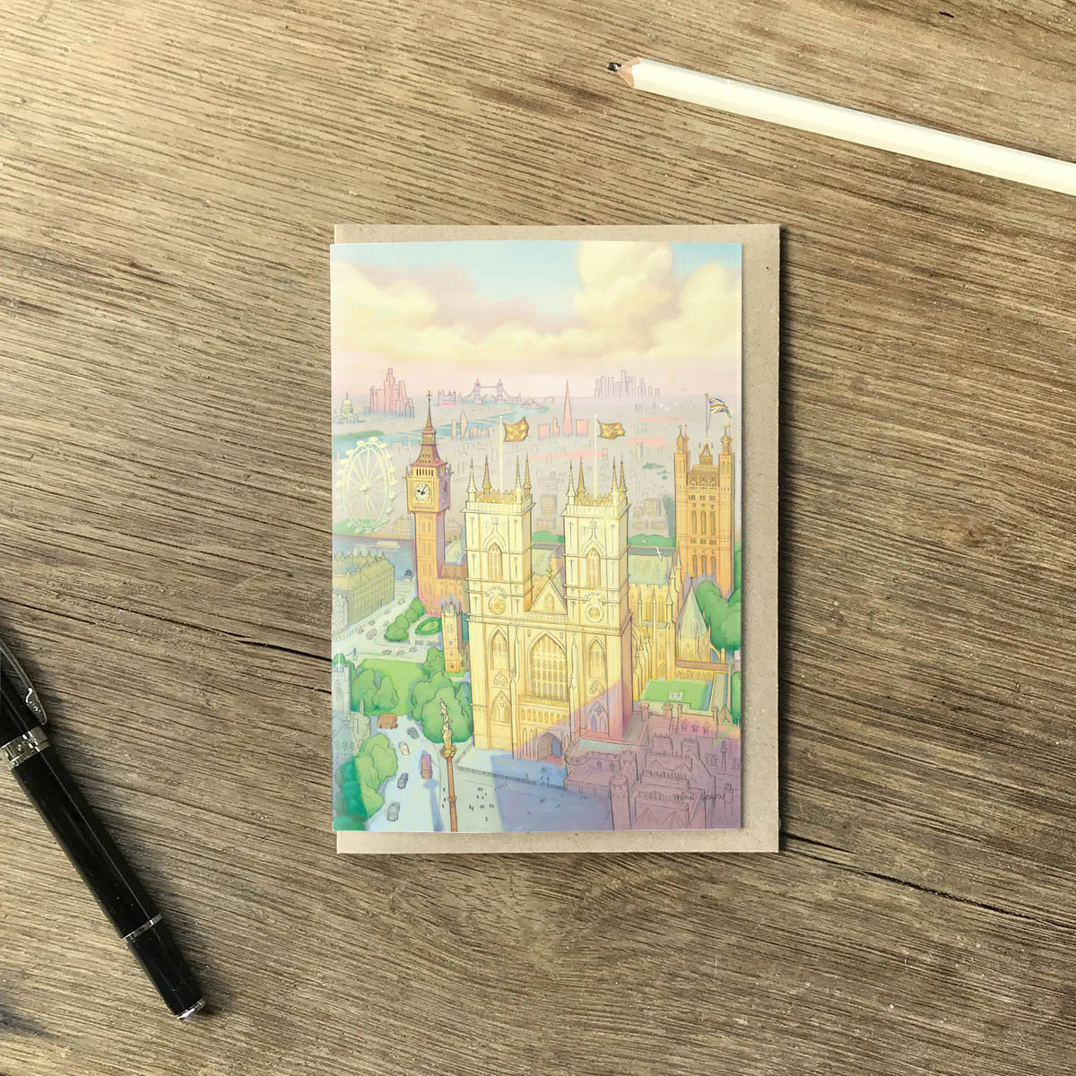 A greetings card featuring a pastel illustration of London landmarks, including Big Ben, Westminster Abbey, and the London Eye, shown with a brown kraft envelope resting on a wooden surface with a pencil and pen.