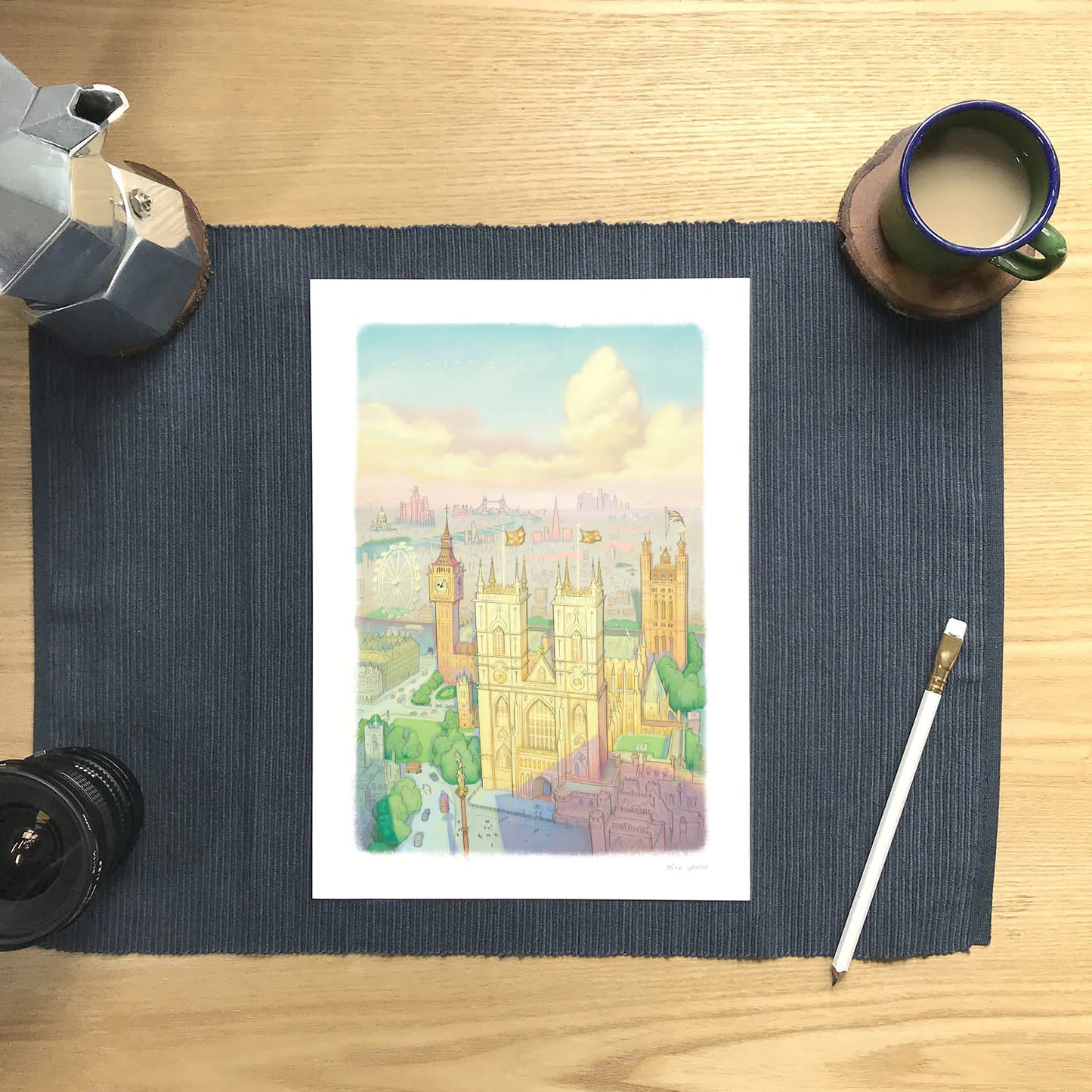 A wall art print of Westminster Abbey in a watercolour illustration style with big ben and the London Eye also shown, resting on a placemat with a coffee and pencil.