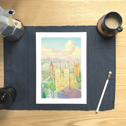 A wall art print of Westminster Abbey in a watercolour illustration style with big ben and the London Eye also shown, resting on a placemat with a coffee and pencil.