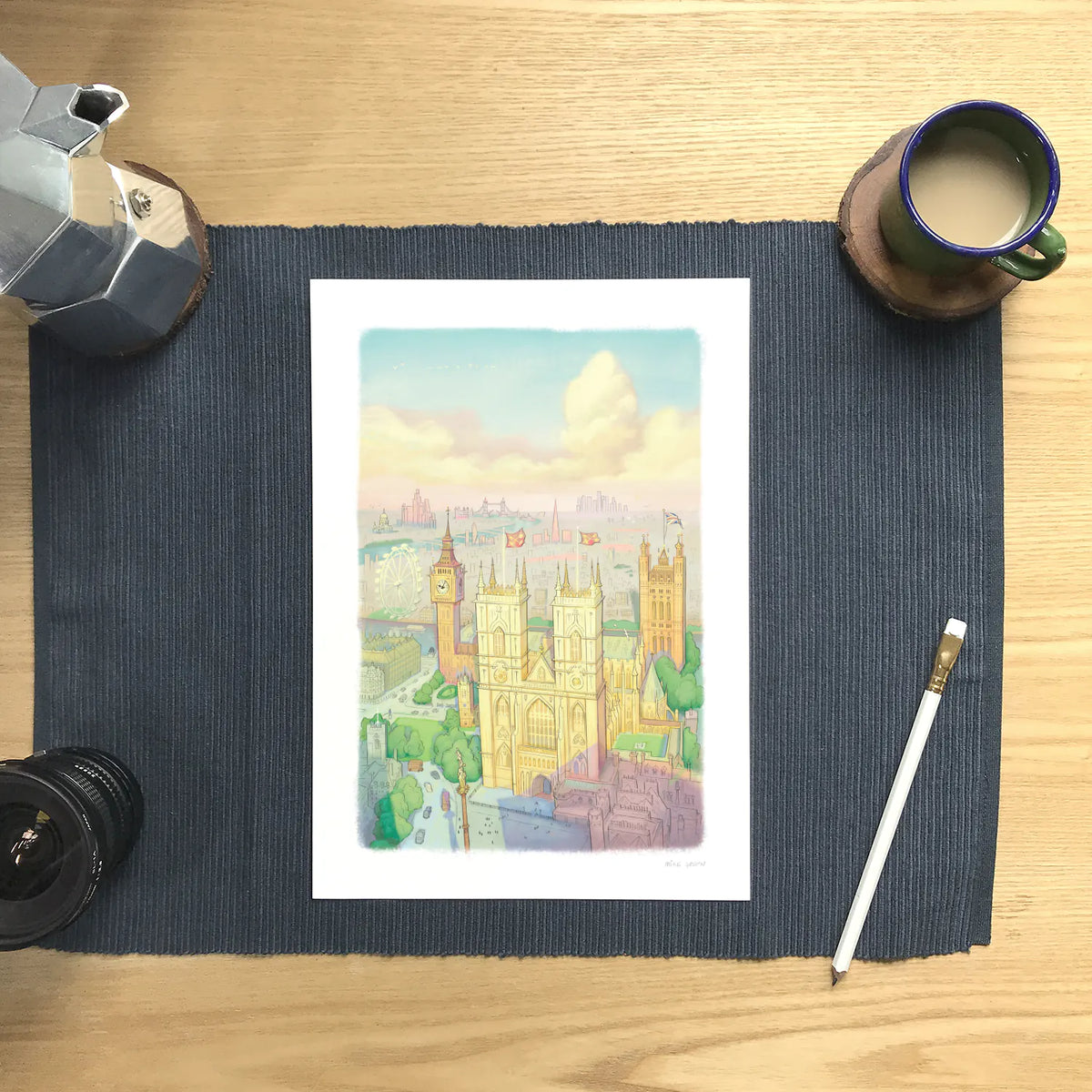 A wall art print of Westminster Abbey in a watercolour illustration style with big ben and the London Eye also shown, resting on a placemat with a coffee and pencil.