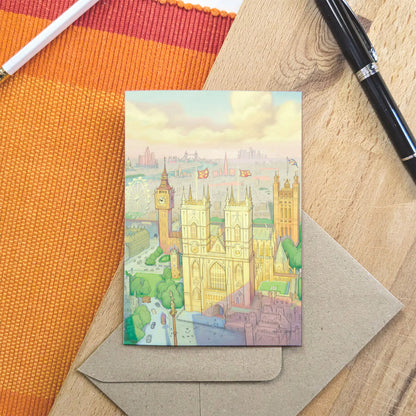 A greetings card featuring a pastel illustration of London landmarks, including Big Ben, Westminster Abbey, and the London Eye, shown with a brown kraft envelope resting on a wooden surface with a pencil and pen.
